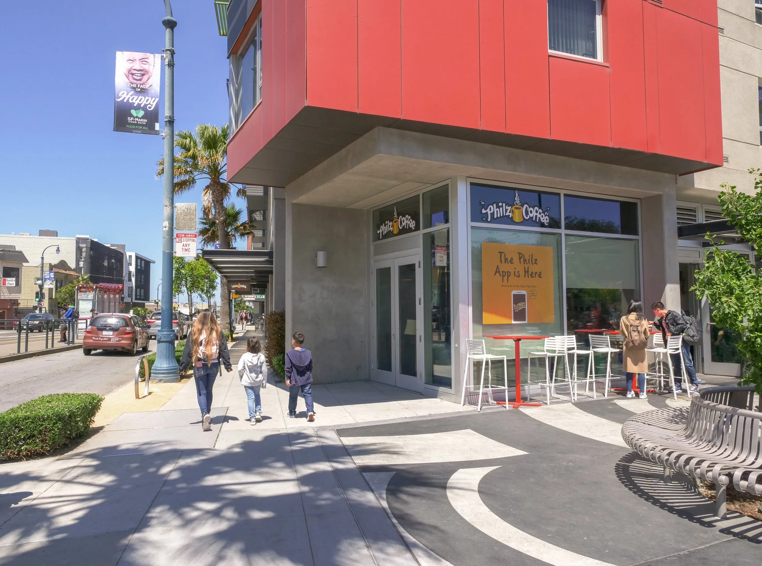 Street scene with a cafe called Philz Coffee on a corner. Three people walking on the sidewalk, two women and a child, and two people standing outside the cafe near white tables and chairs. There are also palm trees, parked cars, and a street lamp wi