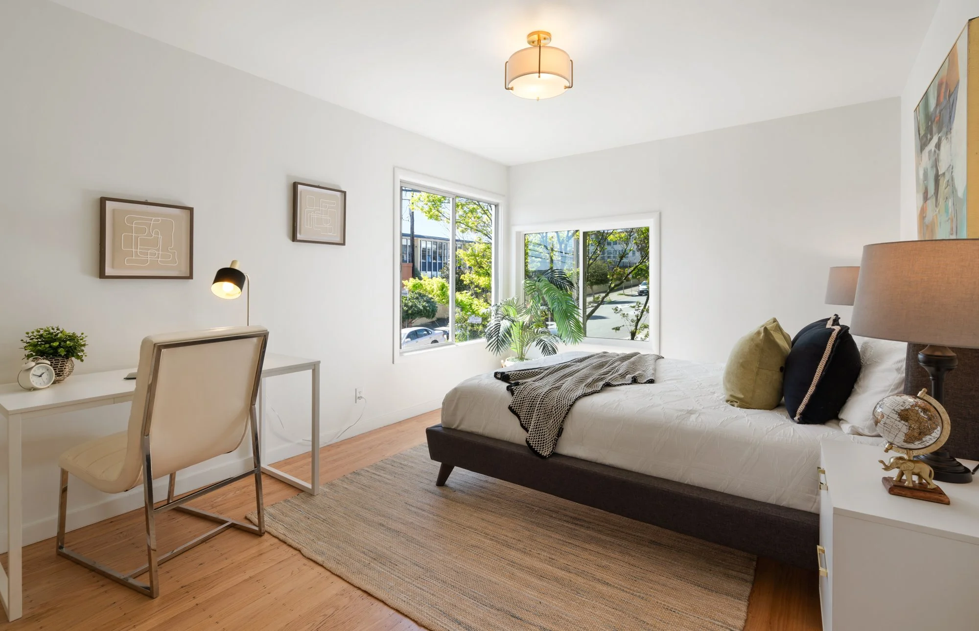 Bright bedroom with white walls, a bed with white bedding and pillows, a window showing outdoor greenery, a small desk with a chair, and a rug on wooden floors.