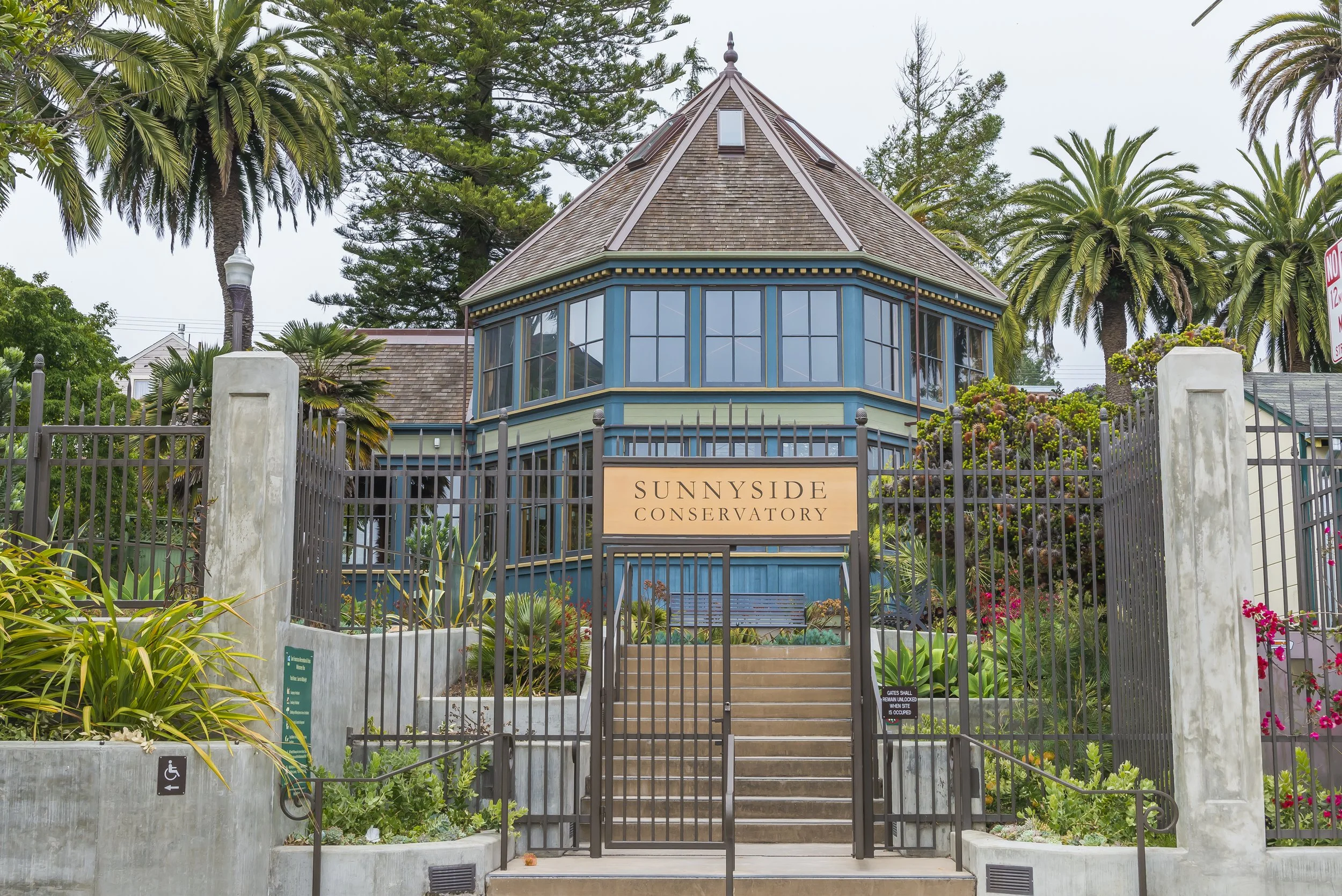 A historic building with a round corner tower, surrounded by palm trees and lush greenery, marked with a sign that reads "Sunnyside Conservatory." The entrance features a gated fence and staircase leading up to the structure.