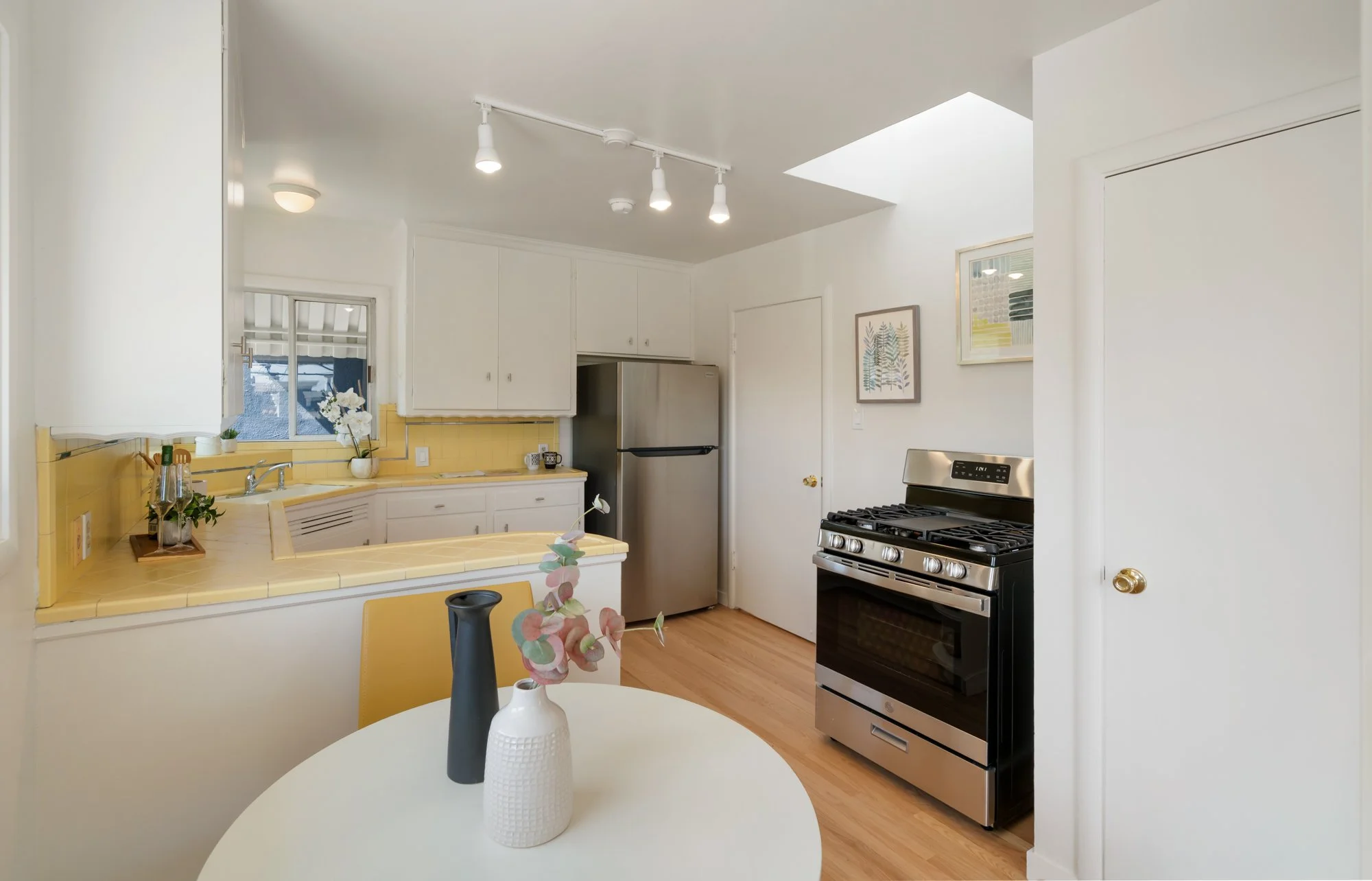 Kitchen with white cabinets, yellow tiled countertop and backsplash, stainless steel refrigerator, black and stainless oven, and decorative vases on a small round dining table.