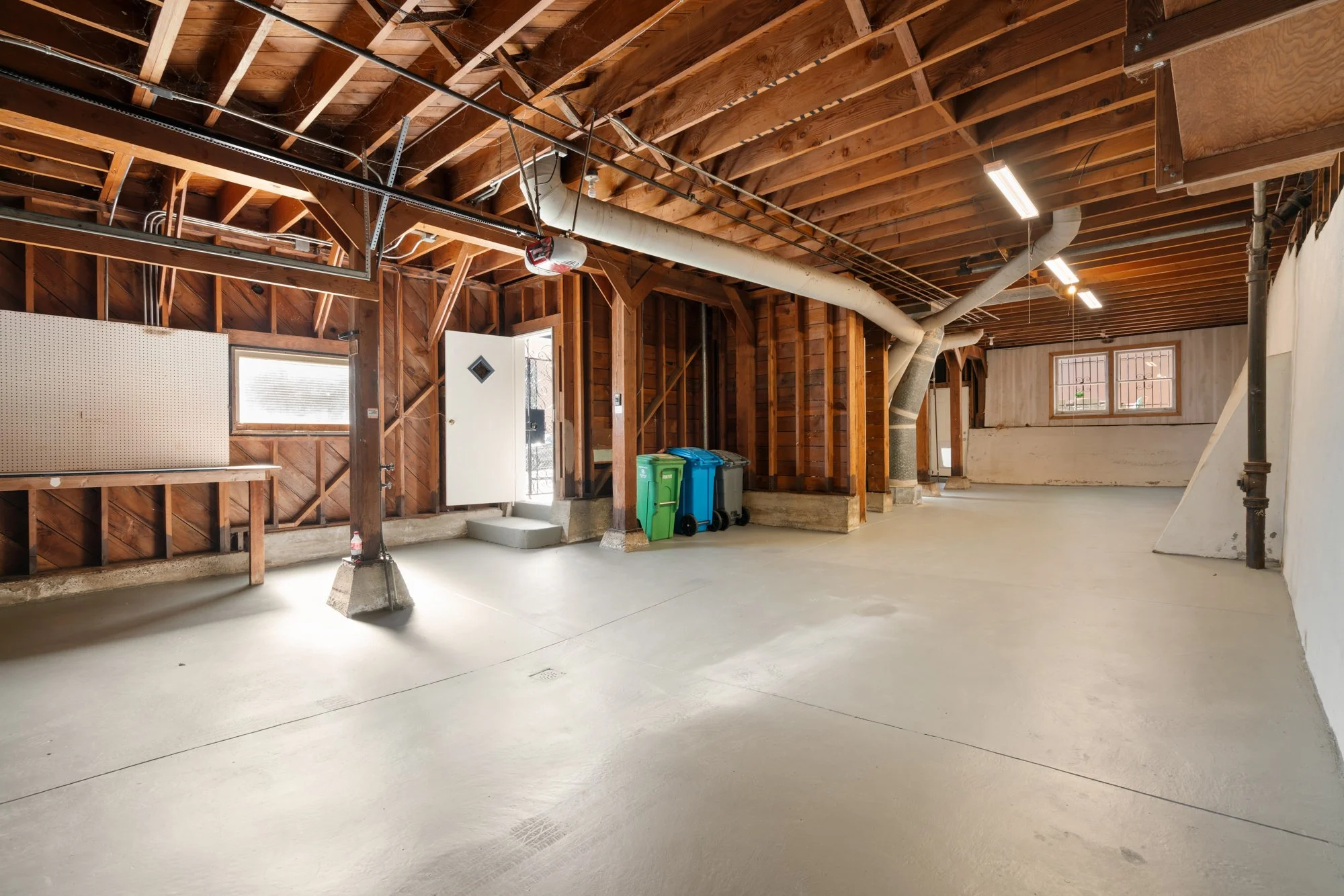 Empty garage with exposed wooden beams, concrete floor, windows, and some trash bins.