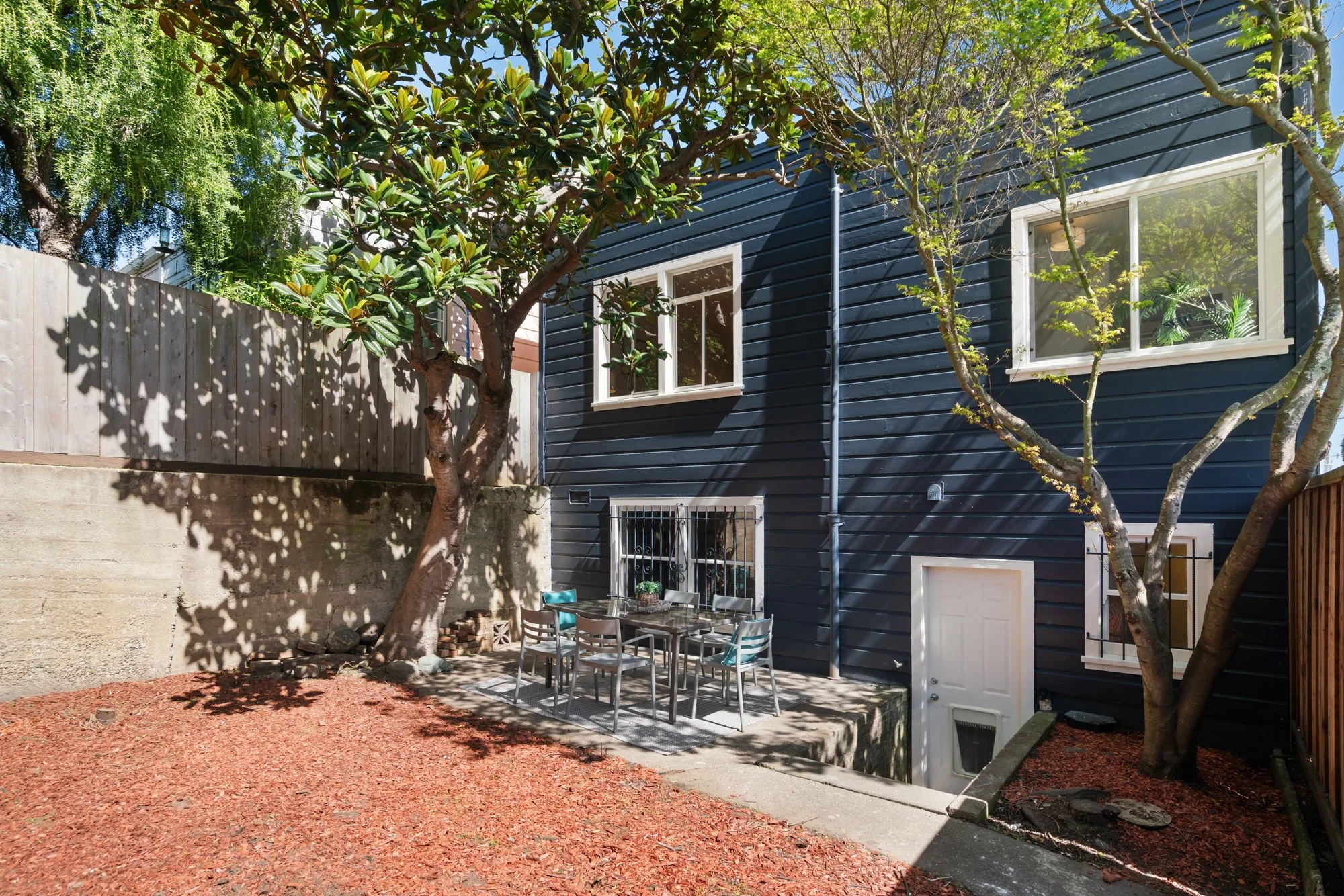 Backyard patio with dining table, chairs, and umbrellas, adjacent to a blue house with white-framed windows and surrounded by trees and a wooden fence.