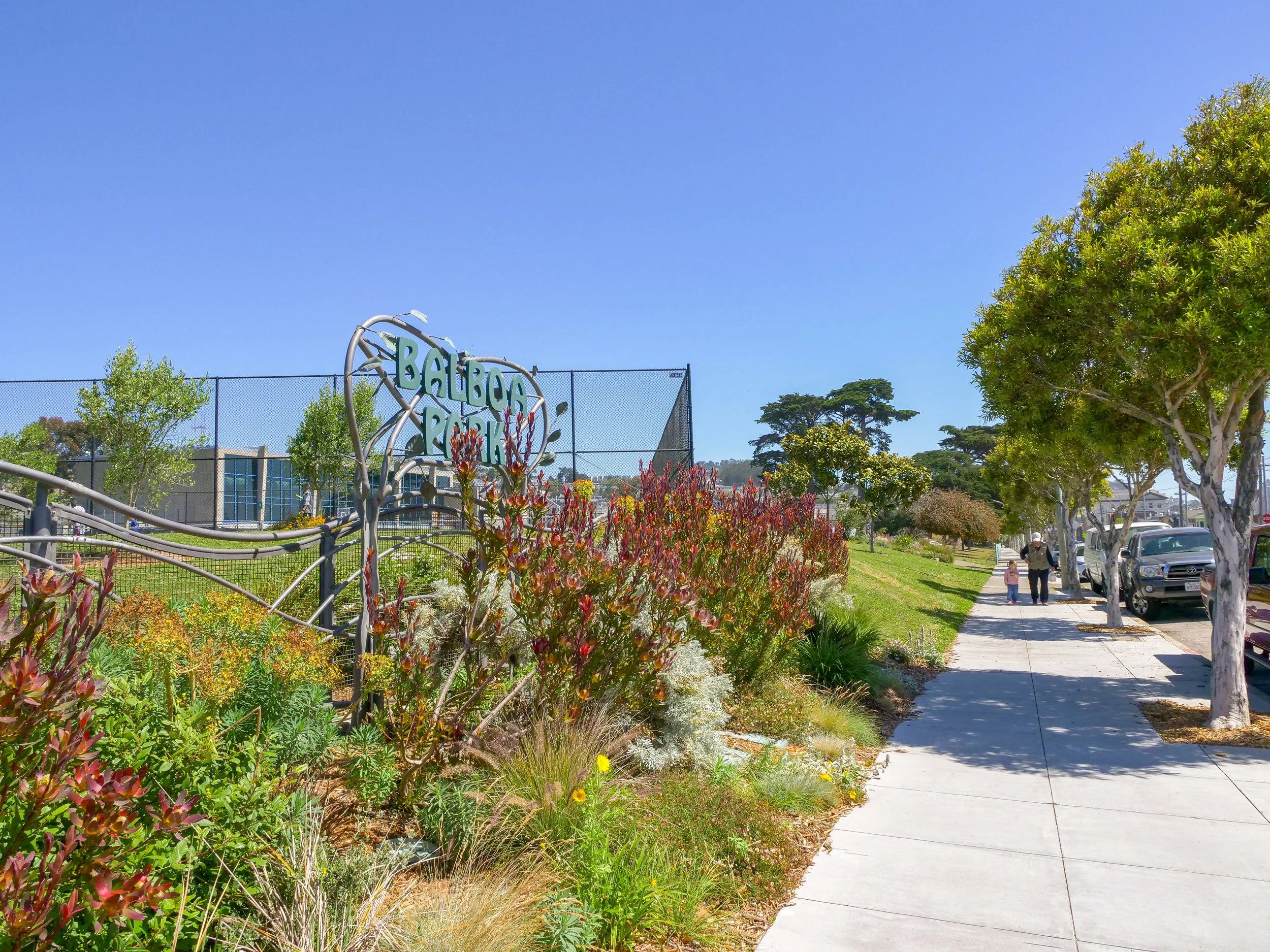 A sunny day at Balboa Park with a sidewalk, trees, and colorful plants next to a tennis court fence.