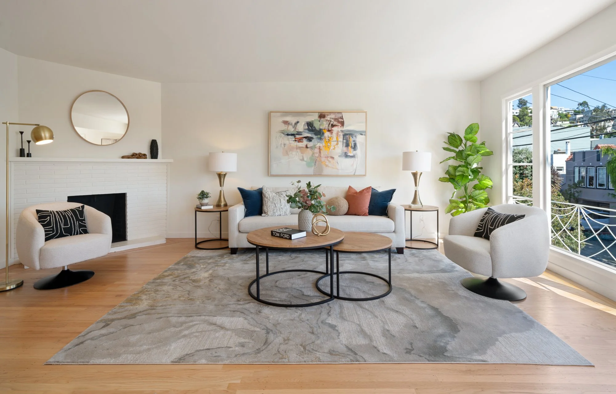 Bright living room with white sofa, two white armchairs, light wood flooring, abstract wall art, potted plants, and large windows with outside view.