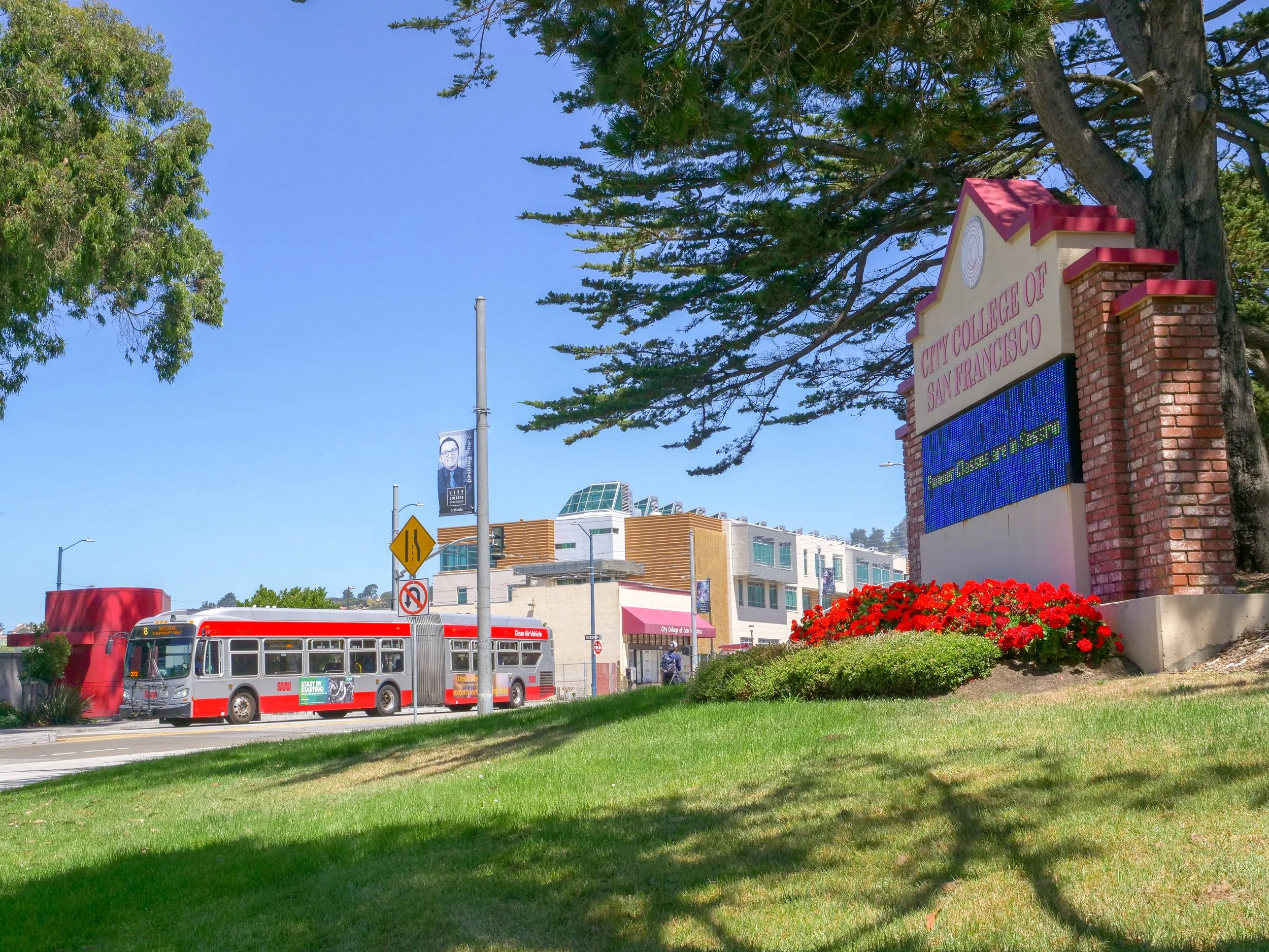 City College of San Francisco campus entrance with a large sign, green lawn, flowers, a bus, and modern buildings in the background under a blue sky.