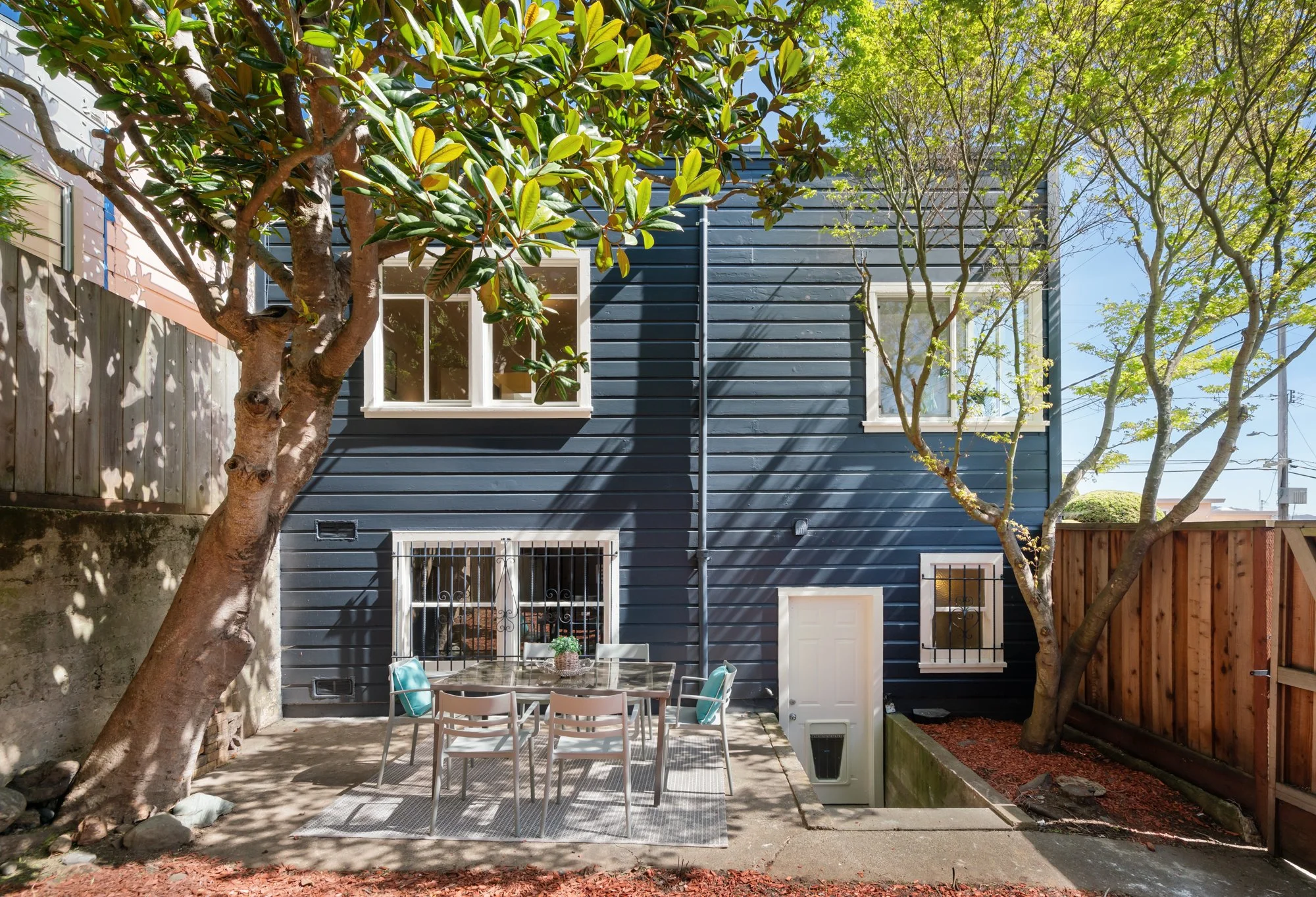 Backyard patio with outdoor table and chairs, two trees, a blue house wall, and a wooden fence.