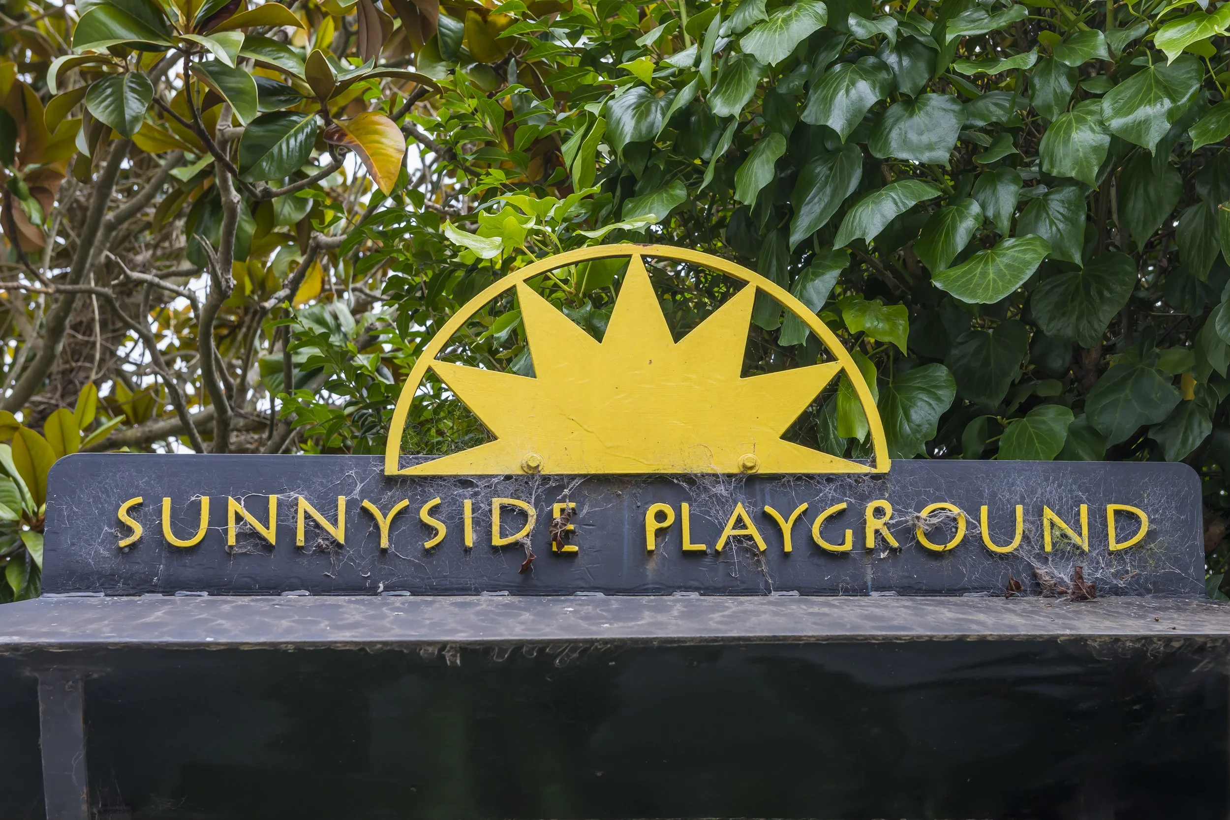 Sign reading 'Sunnyside Playground' with a yellow sun design above, set against a background of green foliage.