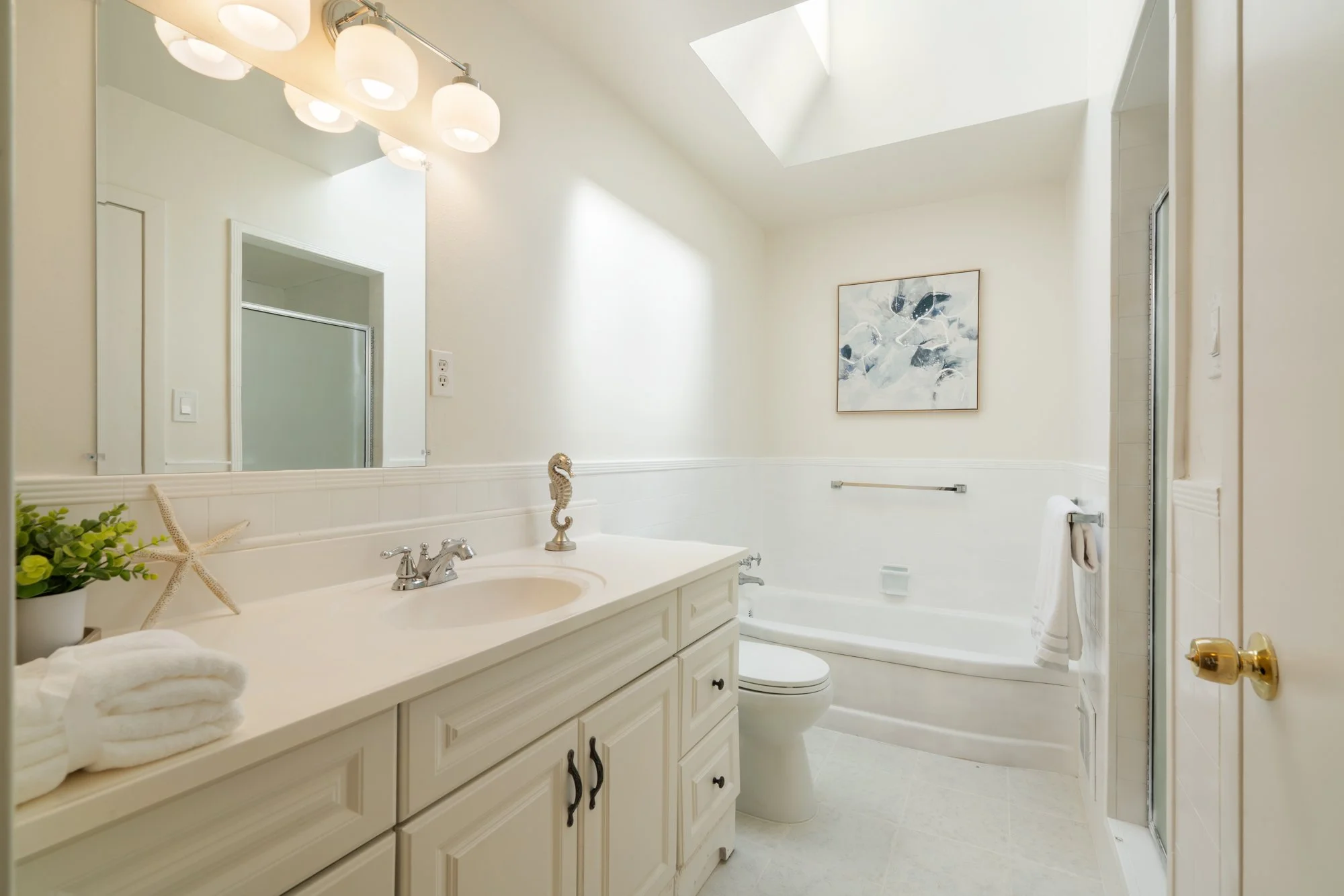 Bright bathroom with white vanity, decorative seahorse, towels, bathtub, and framed artwork with fish, illuminated by skylight.