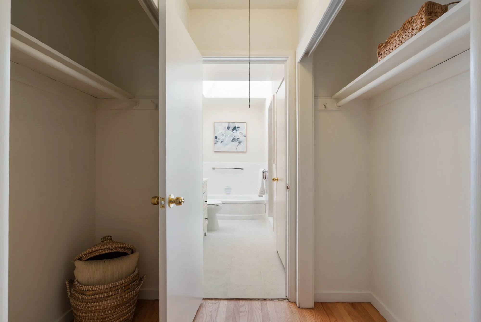 Empty closet with a basket on the floor, leading to a bathroom with a bathtub, toilet, and framed artwork on the wall.