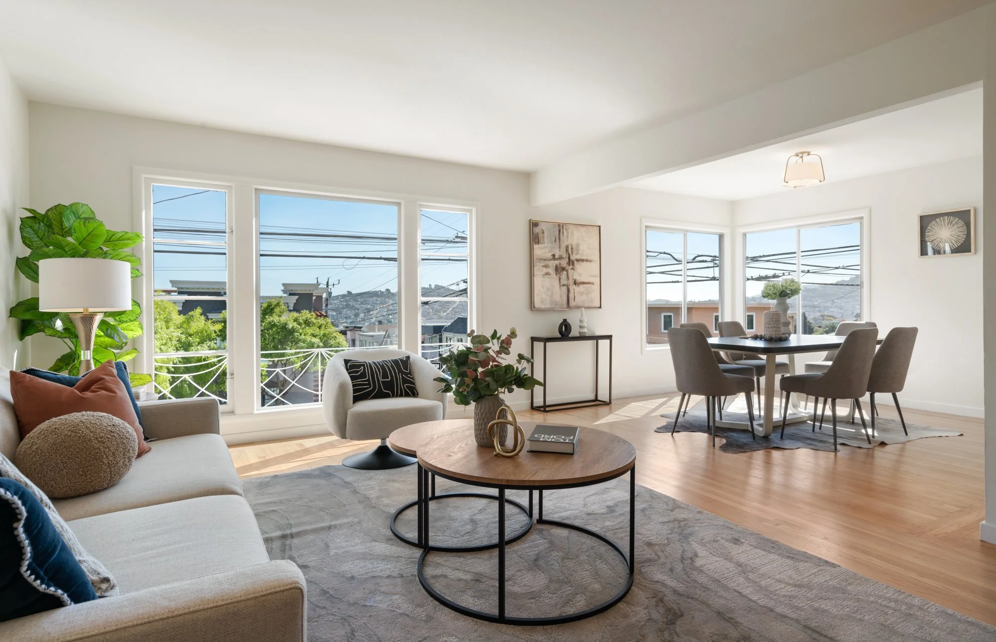 Bright living room and dining area with large windows, white walls, wooden floors, sofa with pillows, round coffee table, armchair, side table, dining table with six chairs, decorative items, and outdoor view of trees and buildings.
