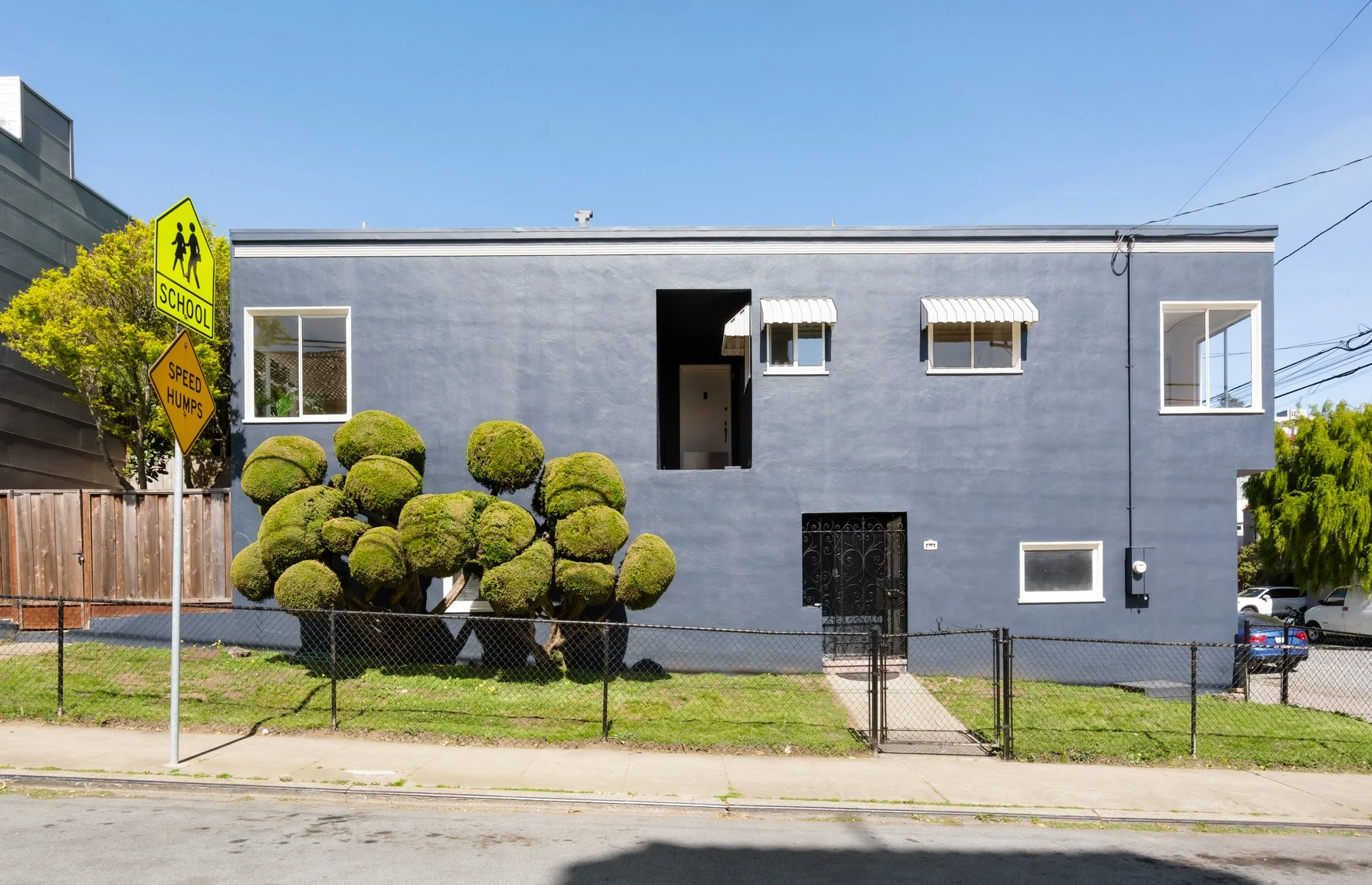 A blue two-story building with white-framed windows and a small, colorful, well-maintained front yard enclosed by a fence. Two street signs indicate a school zone and speed bumps. The sky is clear and blue.