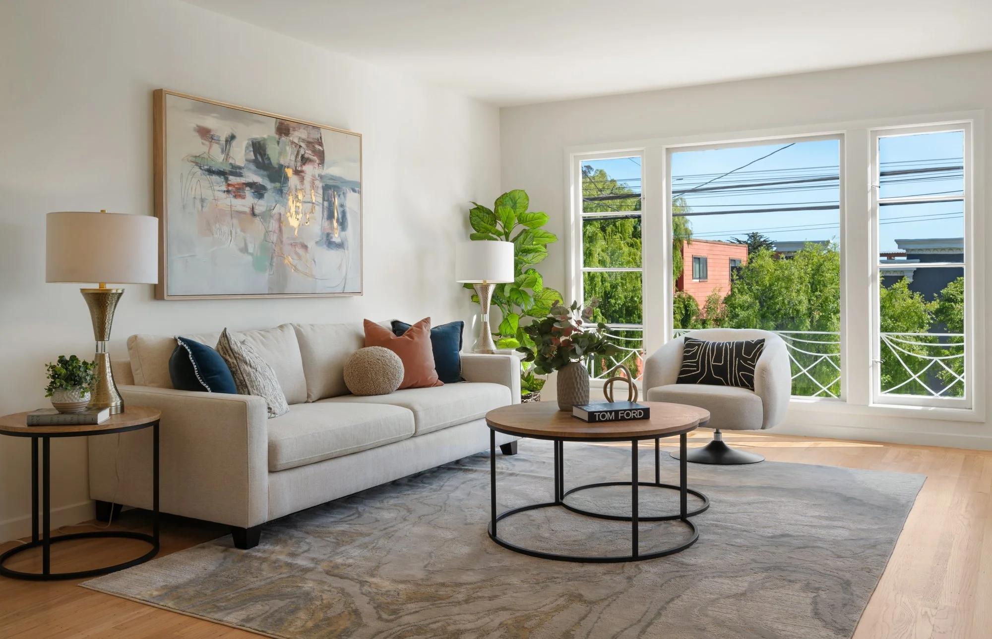 Bright living room with a white sofa, colorful pillows, a round coffee table with a plant and a book, a side table with a lamp, large windows showing greenery outside, hardwood floors, and modern artwork on the wall.