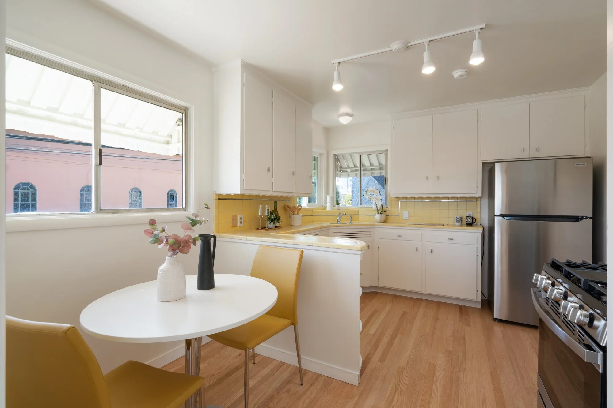 Bright kitchen with white cabinets, yellow tiled backsplash, stainless steel refrigerator, gas stove, and a small dining area with a round white table, two yellow chairs, and a vase with pink flowers.