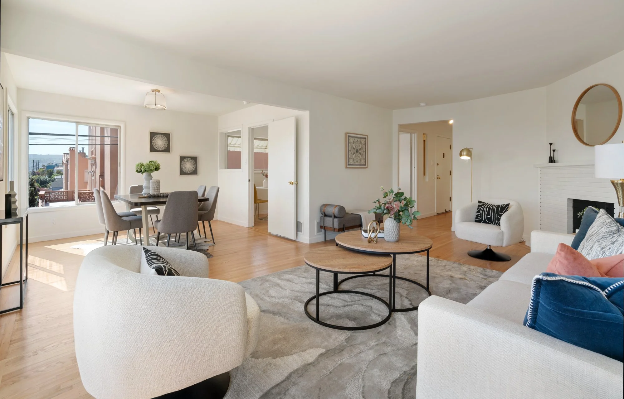 Bright, modern living room with white walls, a white sofa with decorative pillows, a white armchair, wooden coffee tables, a rug, a fireplace, a dining area with a gray table and chairs, large windows with a city view, and decorative wall art.