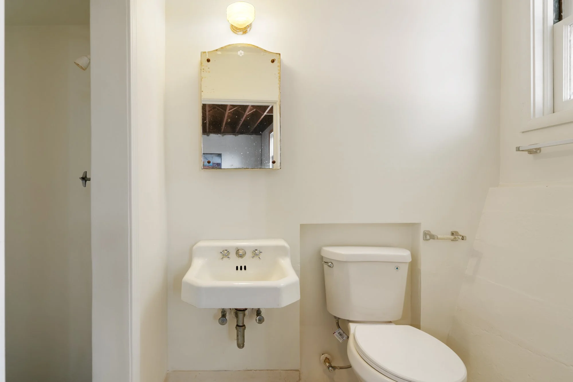 Small bathroom with a white toilet, a white wall-mounted sink with two knobs, a mirror above the sink, a light fixture above the mirror, a window on the right side, and a partially visible door on the left.