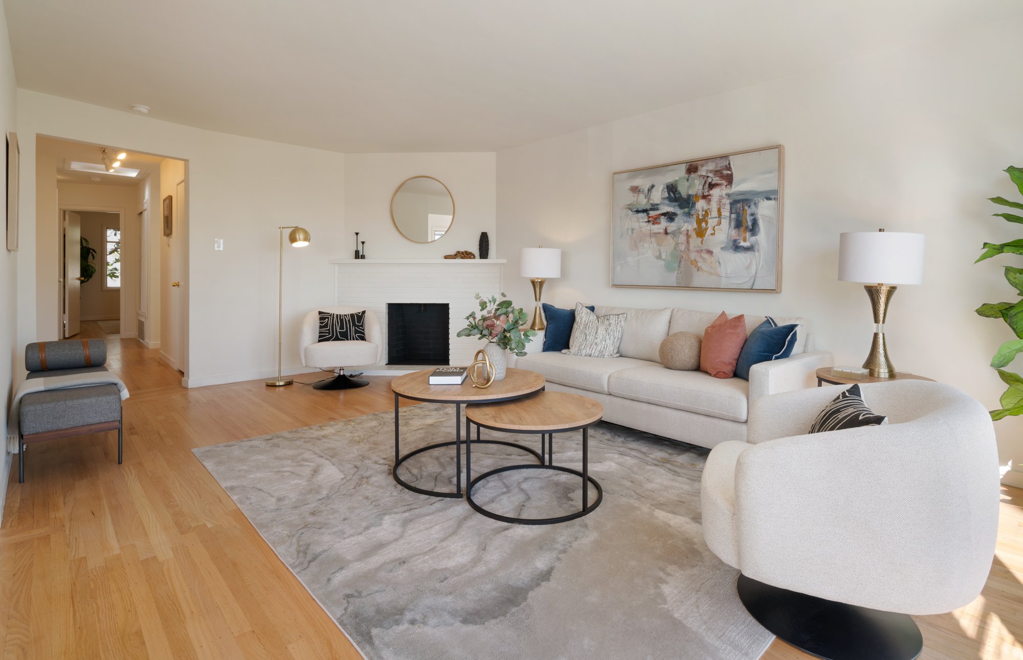 Bright living room with white walls, hardwood floors, a white sofa with colorful pillows, a white armchair, two nested round wooden coffee tables, a fireplace, abstract wall art, and various lamps and decorations.