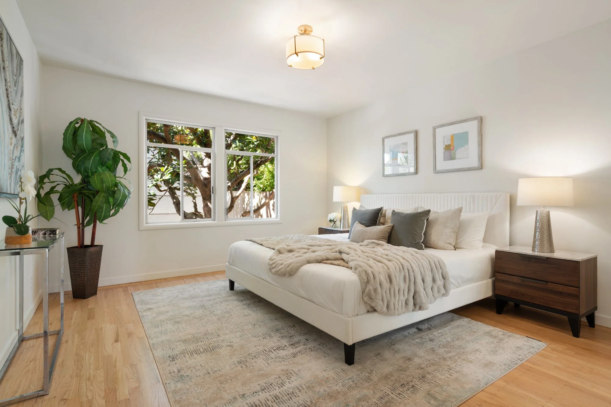 Bright bedroom with a large window showing outside trees, a white bed with multiple pillows, a beige fur throw, two bedside tables with lamps, a large potted plant, artwork on the wall, and a patterned area rug on wooden floors.