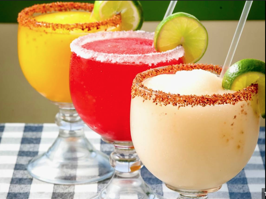 Three colorful frozen drinks in margarita glasses with lime wedges and salted rims, arranged on a checkered table.