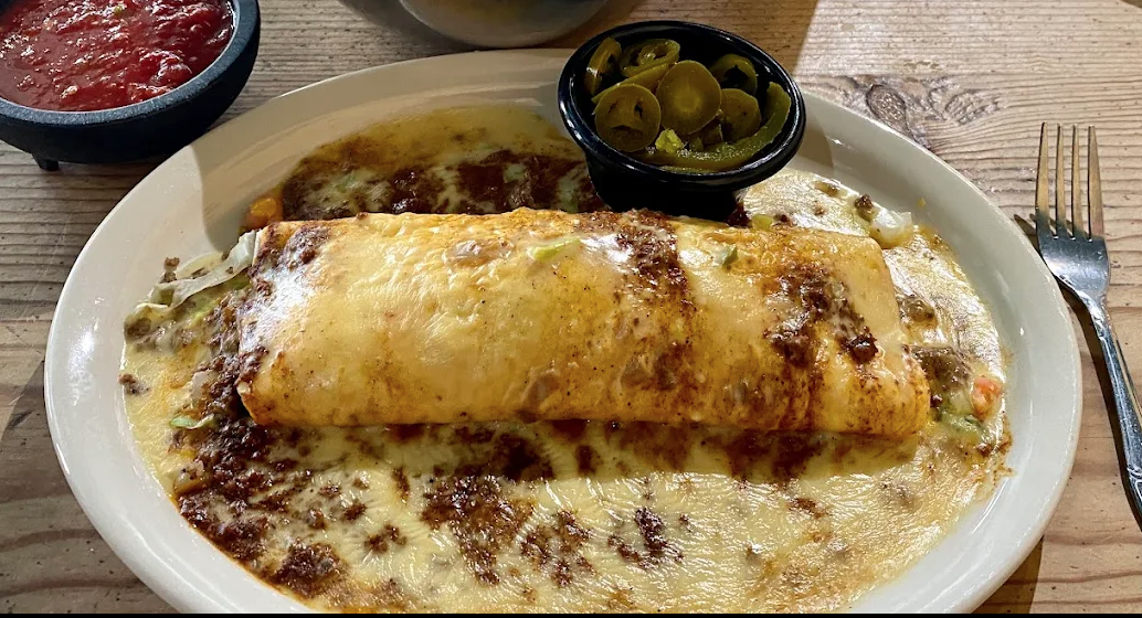 Plate with an enchilada covered in melted cheese, served with a side of jalapeños and a small dish of red salsa.