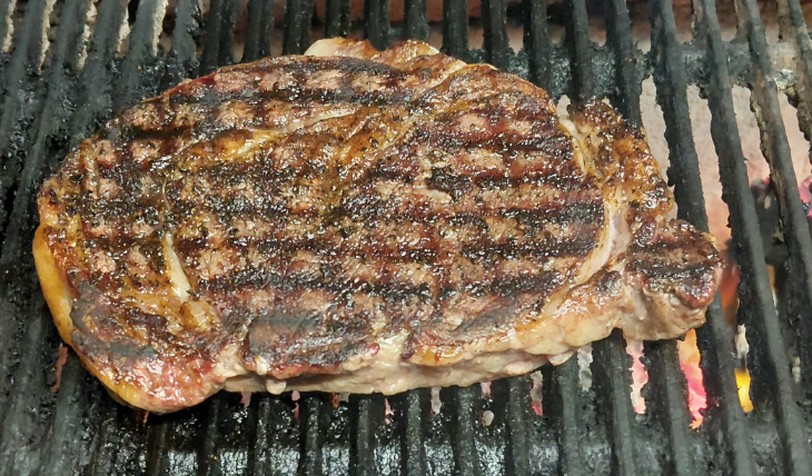 A beef steak cooking on a grill with grill marks visible.