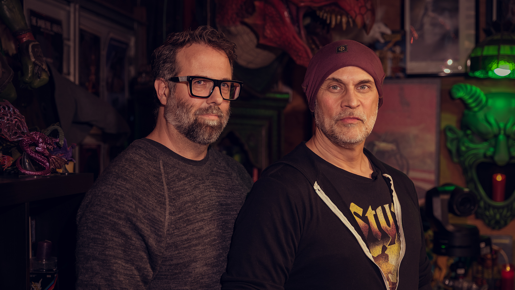 Two middle-aged men with beards and serious expressions, standing in a room decorated with colorful artwork and collectibles, including a large dragon head sculpture and neon green lighting in the background.