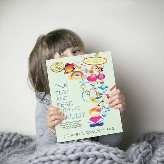 Child holding a colorful children's book titled 'Talk, Play, and Read with Me Daddy' in a cozy setting with a gray blanket.