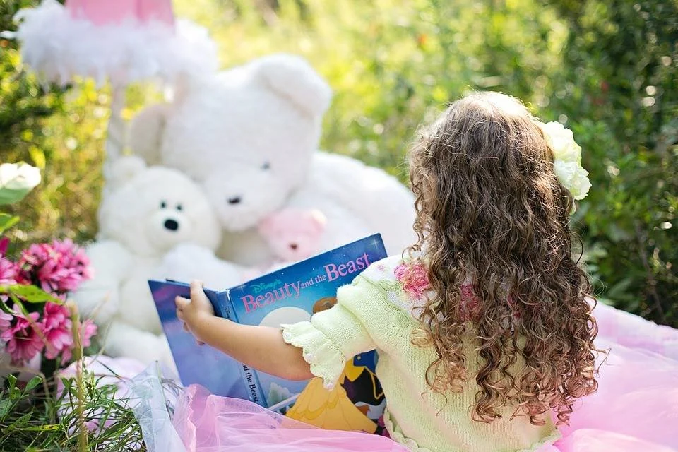 A young child sits outside in a garden reading a Beauty and the Beast book to large white teddy bears, surrounded by flowers and soft pink fabric.