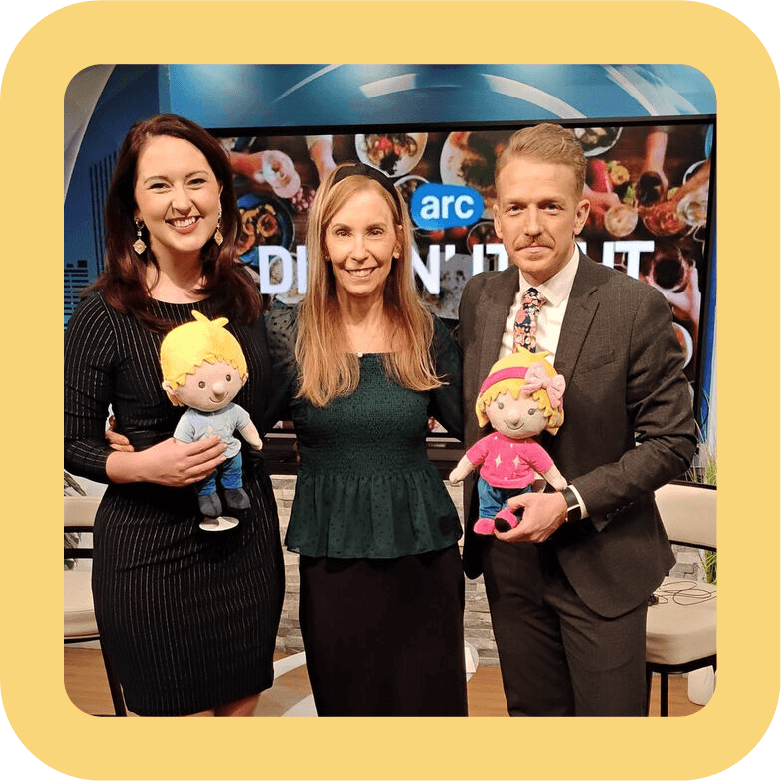 Three people standing indoors, two women and one man, each holding a plush doll. A TV screen behind them displays the logo for ABC and the word 'DIALOGUE'. The setting appears to be a television studio with chairs and a backdrop.