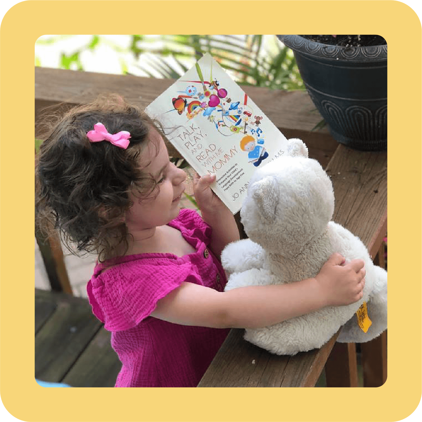 A young girl with curly hair, wearing a pink dress and pink bow, reading a colorful children's book to a white teddy bear, sitting at a wooden table outdoors.