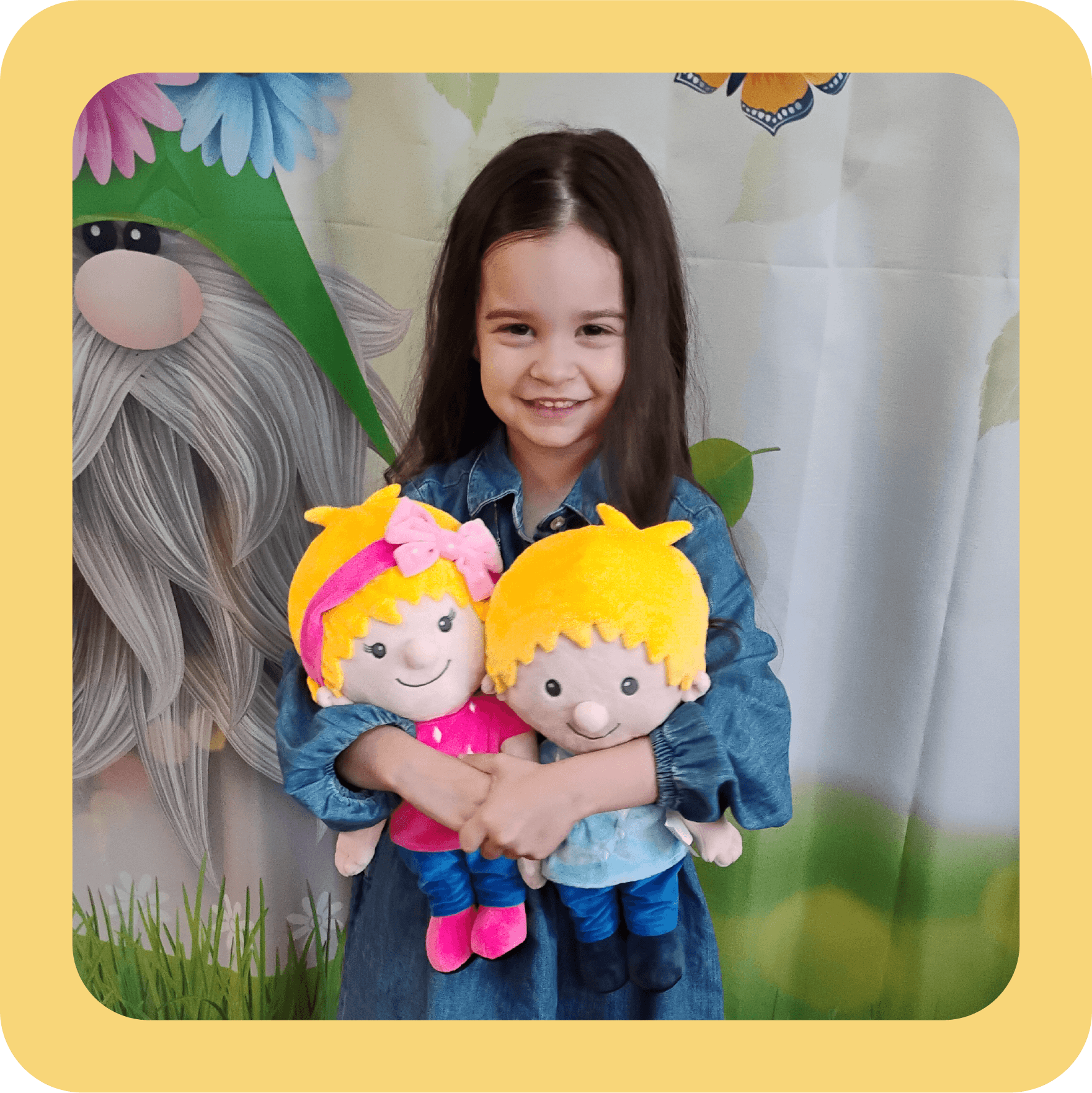 A young girl with long dark hair smiling and holding two plush dolls, Ruby and Louie, with blonde hair, dressed in pink and blue. She is standing in front of a colorful backdrop with a gnome and floral decorations.