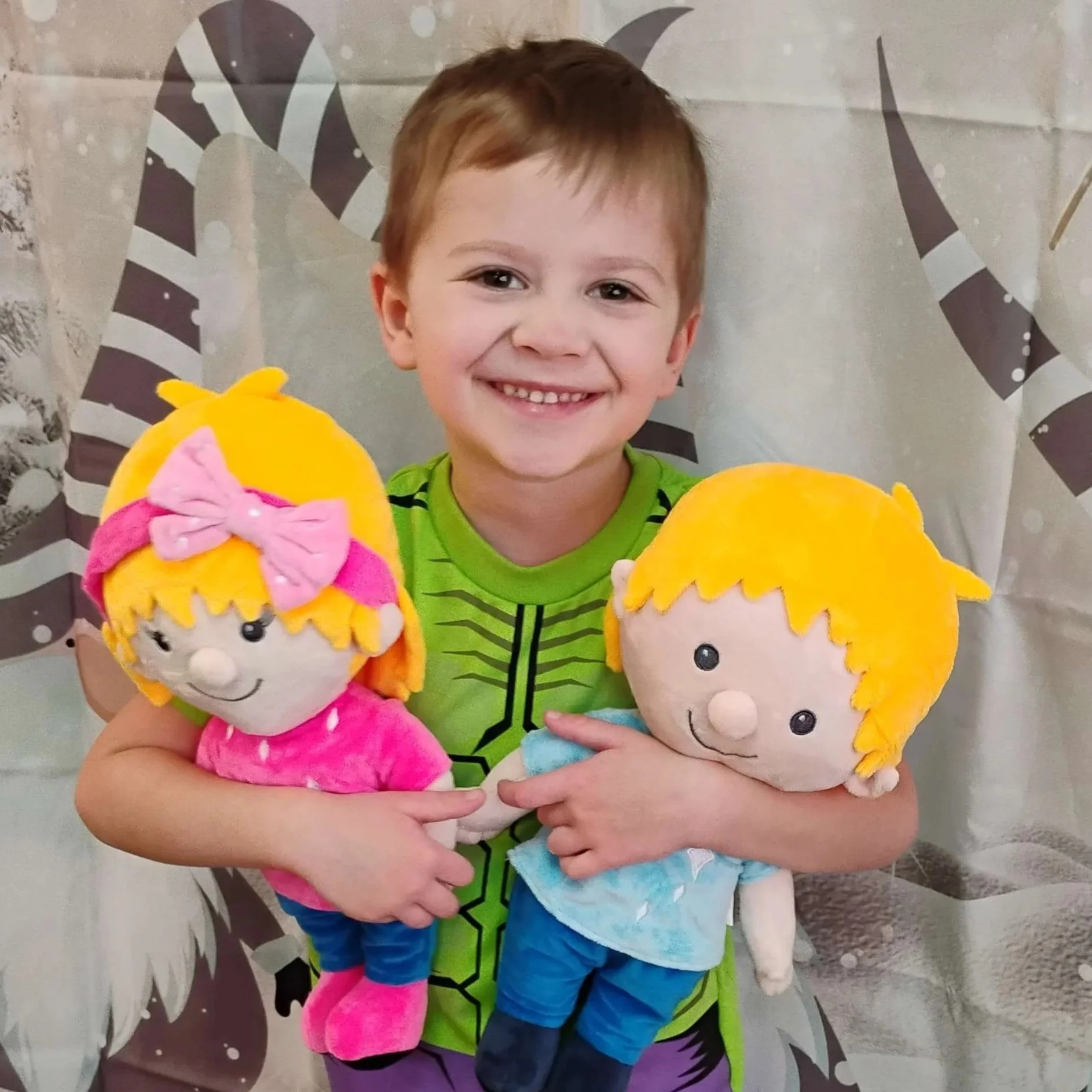 A young boy with short brown hair and a bright green shirt, smiling, holding two plush dolls with blond hair and round faces, one dressed in pink with a bow, the other in light blue. They are standing in front of a gray background with cartoon-like graphic elements.