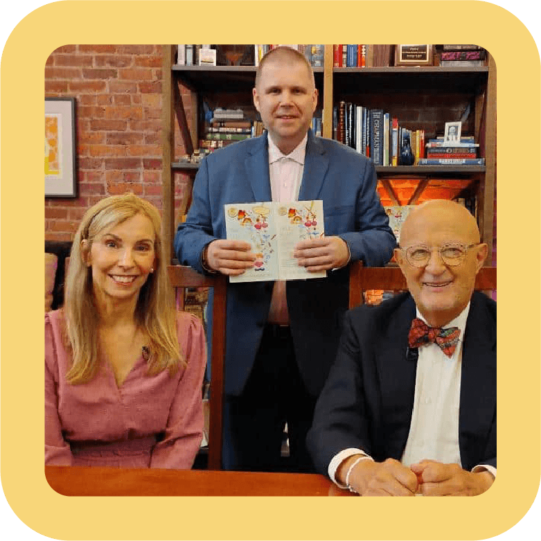 Three people sitting and standing in a room with bookshelves; a woman in pink on the left, a man in a suit with a bow tie on the right, and a man standing in the middle holding a book, with a brick wall and bookshelves in the background.