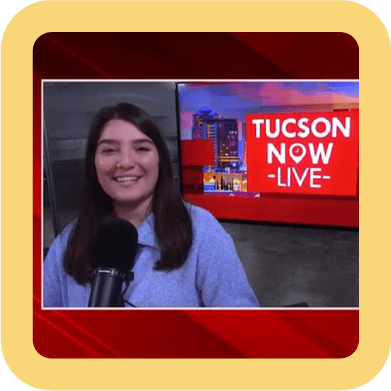 A woman with dark hair and a blue sweater smiling on a news set, with a microphone in front of her and a red and blue background that says 'TUCSON NOW - LIVE'.