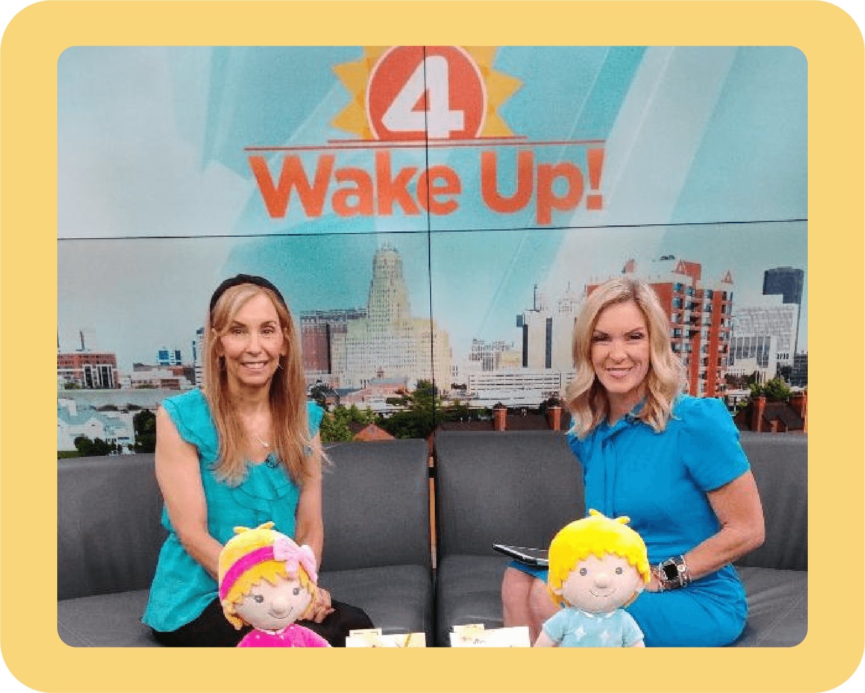 Two women sitting on a gray couch with plush toys in front of them, a cityscape background on TV, and a graphic that says '4 Wake Up!'
