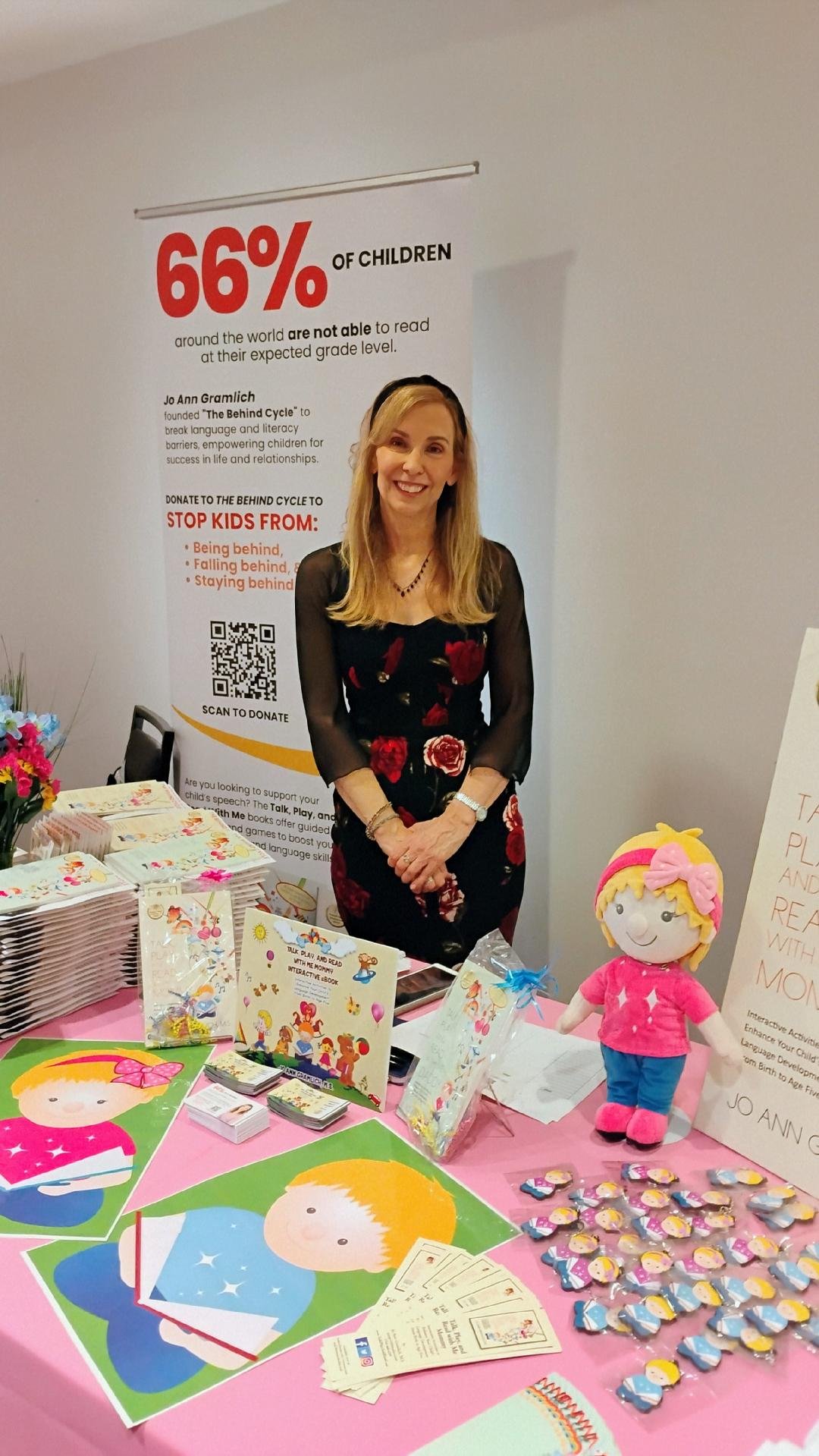 A woman, Jo Ann Gramlich, standing behind a table with children's plush toys, Ruby and Louie, and educational materials, at a booth supporting literacy and language development, with a large poster about childhood literacy behind her.