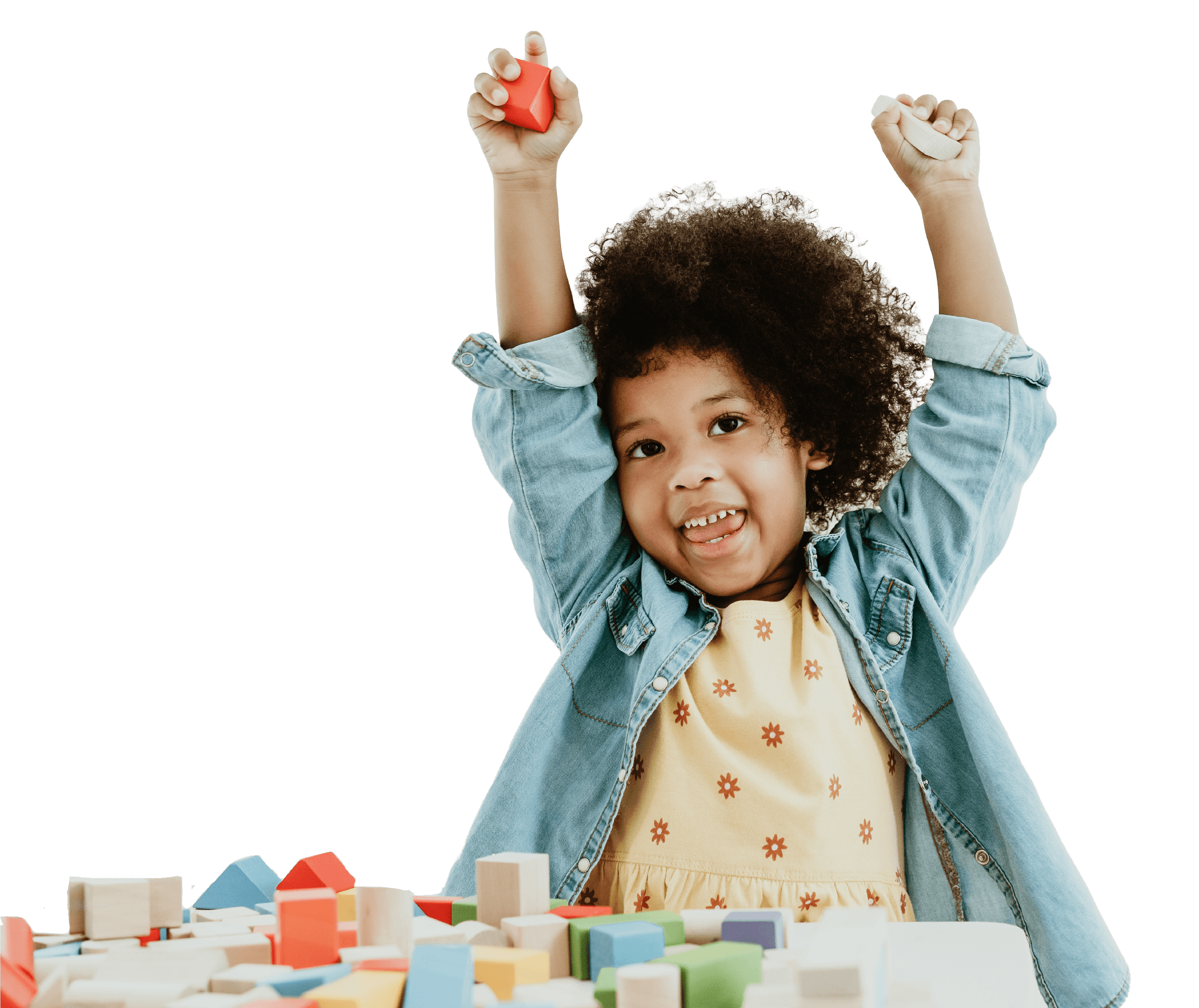 Young girl with curly hair wearing a denim jacket and yellow dress, smiling and raising her arms, surrounded by colorful wooden blocks.