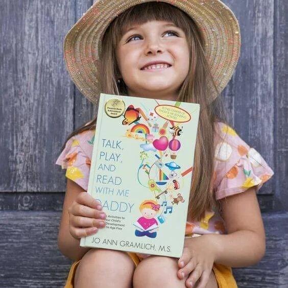 A young girl wearing a straw hat smiling and holding a book titled 'Talk, Play, and Read with Me Daddy' by Jo Ann Gramlich, M.S. standing against a wooden background.