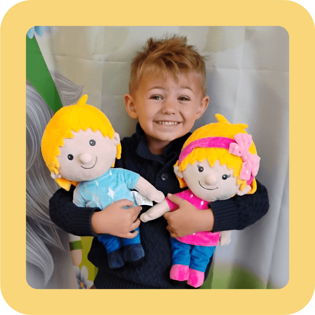 A young boy with a big smile holding two plush dolls, Ruby and Louie, with yellow hair, one dressed in blue and the other in pink.