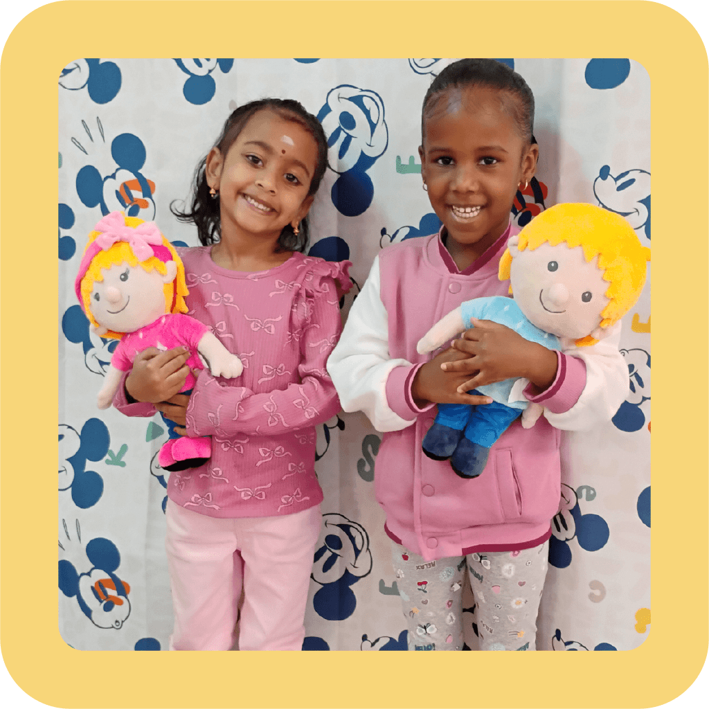 Two young girls smiling and holding plush dolls, Ruby and Louie, standing in front of a Disney-themed backdrop.