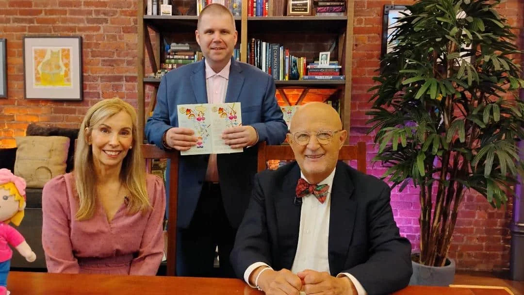 Three people sitting and standing together in a cozy room with bookshelves and brick walls. The man seated in front wears glasses, a bow tie, and a suit. The woman sitting to his left has long blonde hair and a pink blouse. The man standing behind them holds a colorful book and wears a blue blazer.