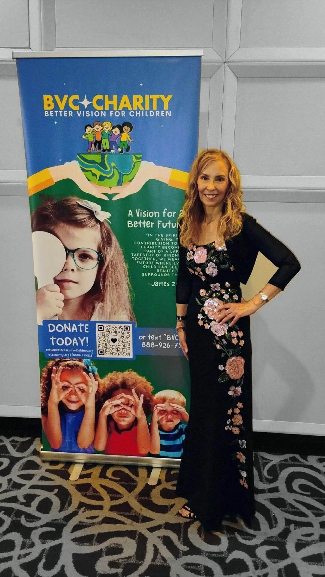 A woman, Jo Ann Gramlich, standing next to a banner for BVC Charity with children and colorful graphics, at an indoor event.