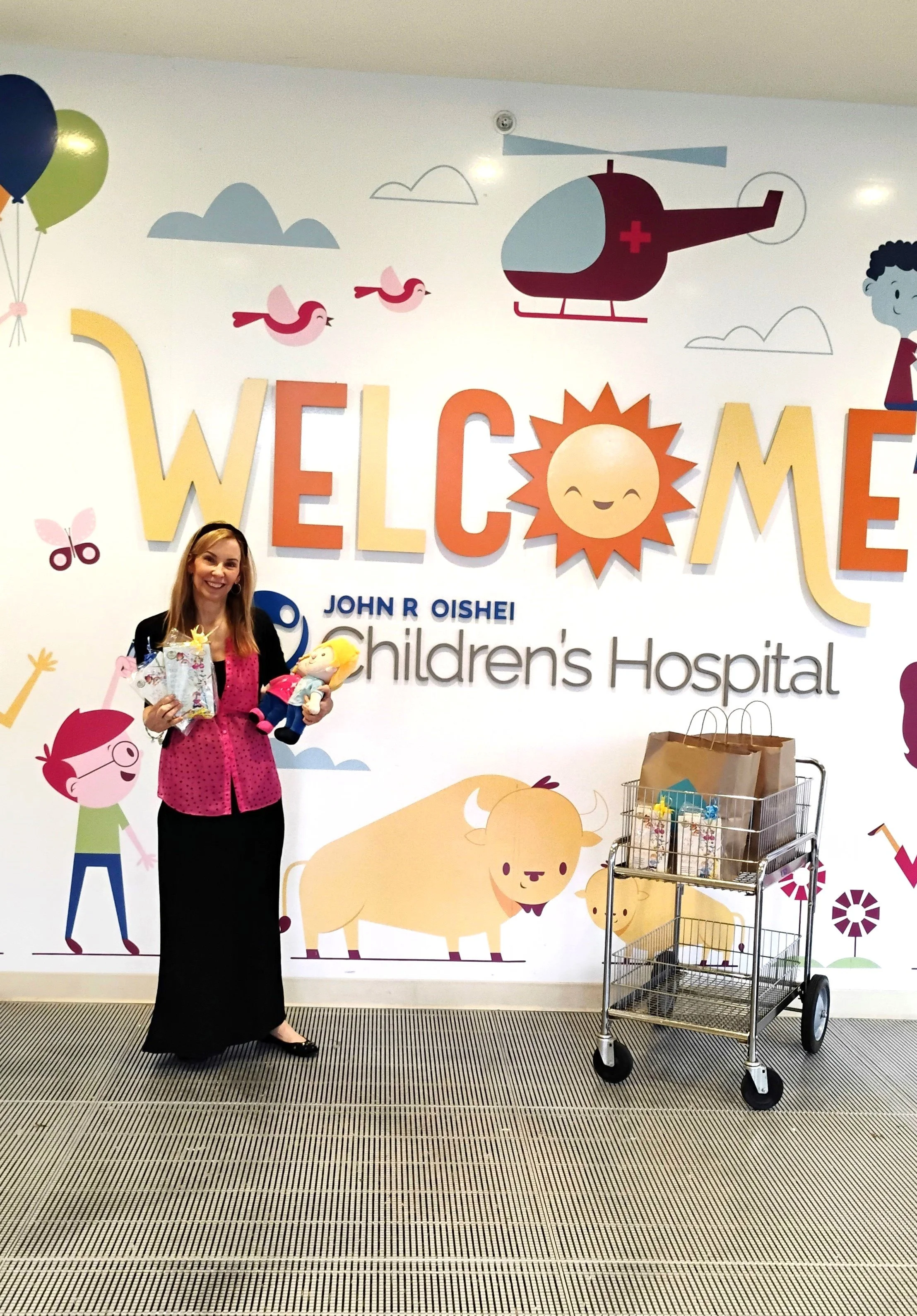 A woman standing in front of a children's hospital mural, holding toys and gifts, with a cart nearby loaded with presents. The mural features colorful cartoon animals, a helicopter, and cheerful characters with a welcoming message.