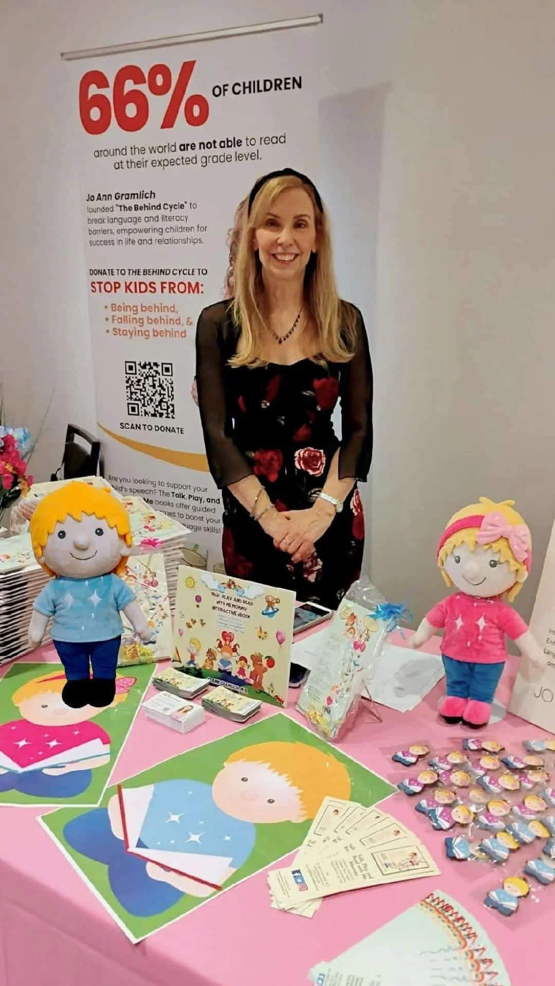 A woman, Jo Ann Gramlich, standing behind a table with children's plush toys, Ruby and Louie, and educational materials, at a booth supporting literacy and language development, with a large poster about childhood literacy behind her.