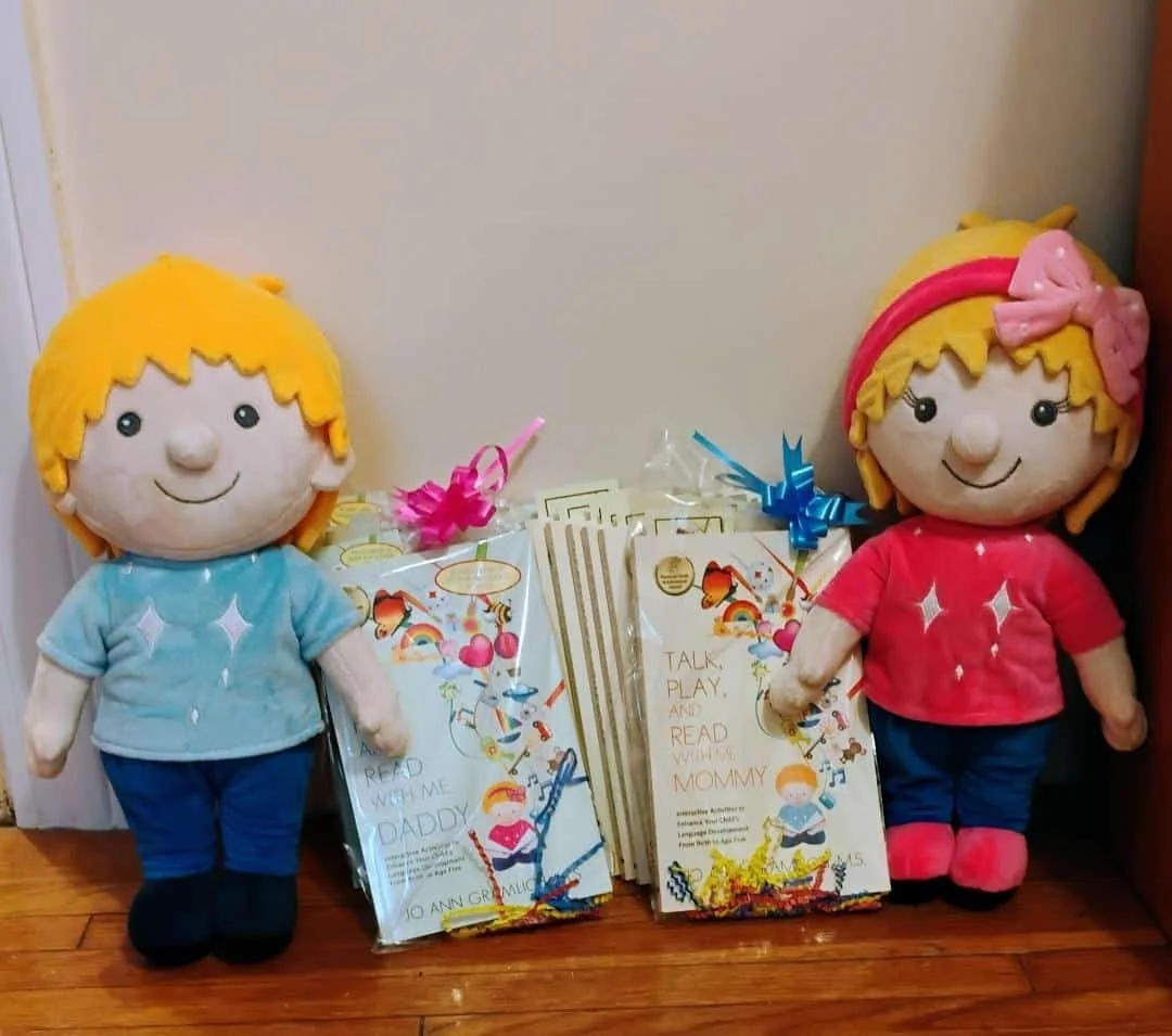 Two plush dolls, one boy with yellow hair and a blue shirt, and one girl with yellow hair, pink headband, and pink shirt, are standing next to a stack of children's books about reading with parents.