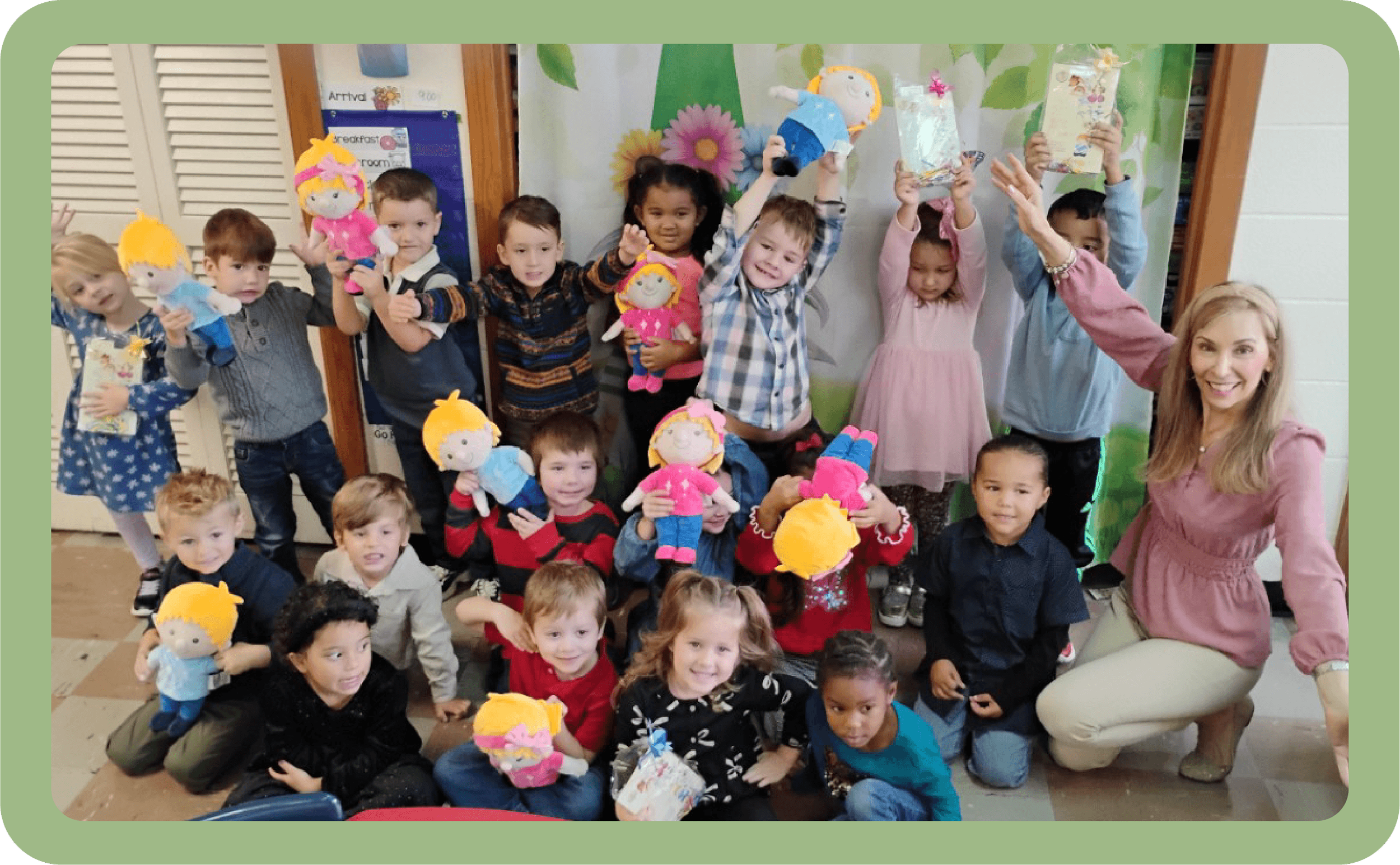 Group of children and a woman in a classroom, holding and displaying plush dolls, smiling, with a floral backdrop in the background.