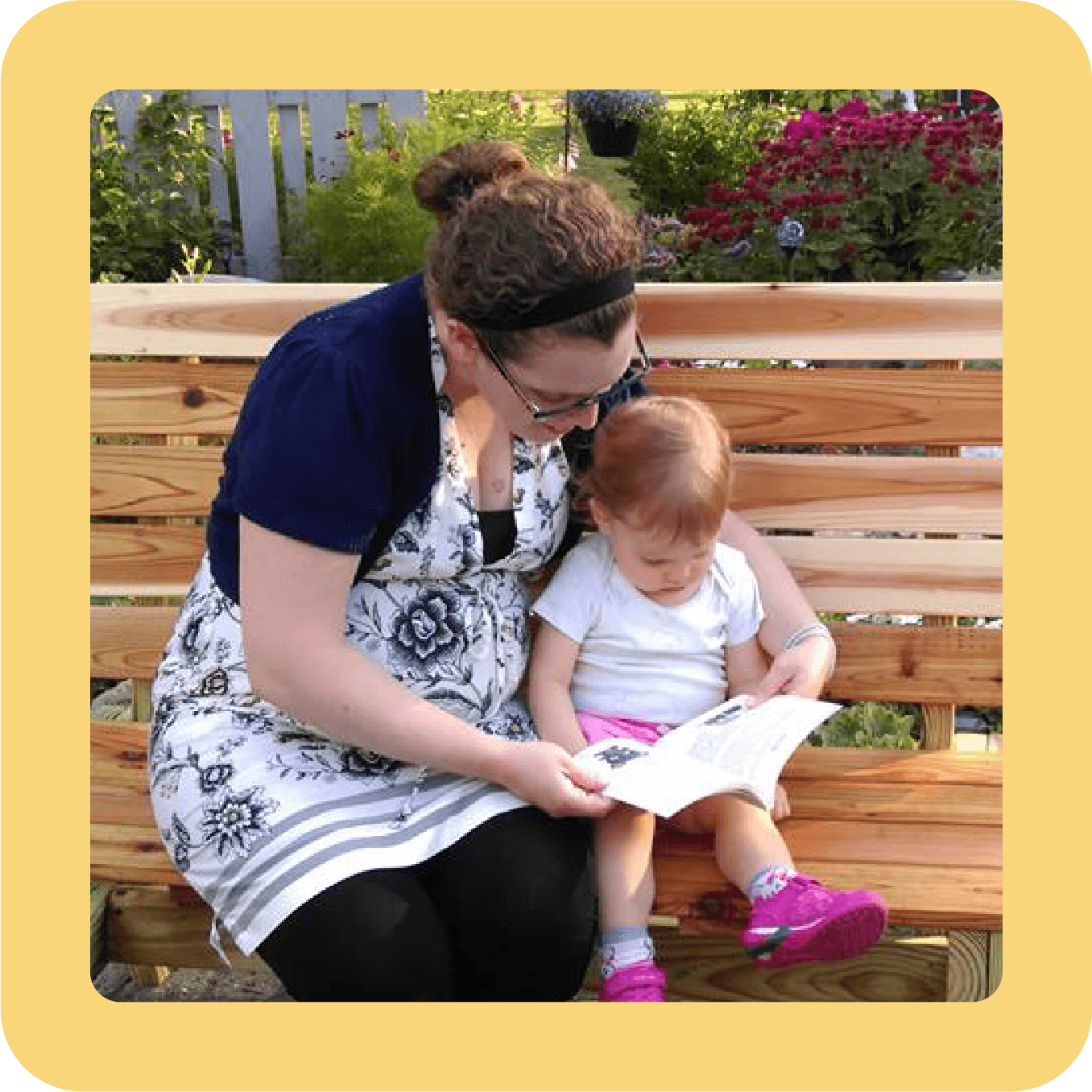 A woman and a young girl sitting on a wooden bench outdoors, reading a book together. Flowers and greenery are in the background.