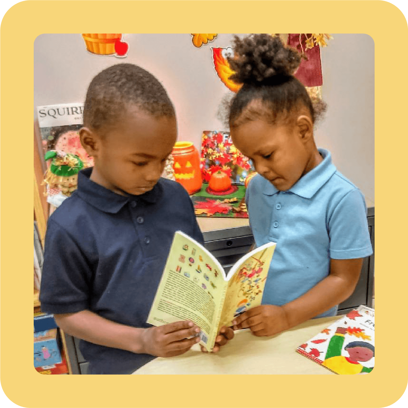 Two young children, a boy and a girl, are looking at a book together in a classroom with Thanksgiving decorations in the background.