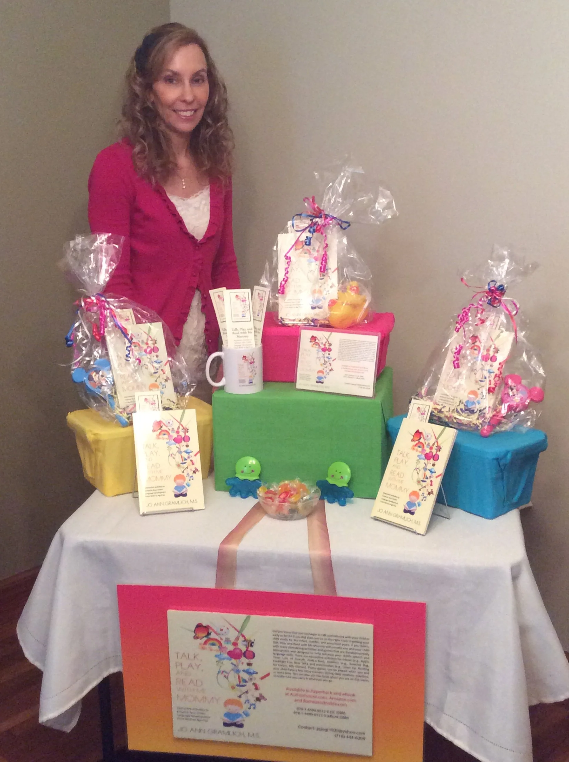 Woman, Jo Ann Gramlich, smiling at a table with colorful gift baskets, informational pamphlets, and a large pink sign promoting reading, talking, and playing with children.