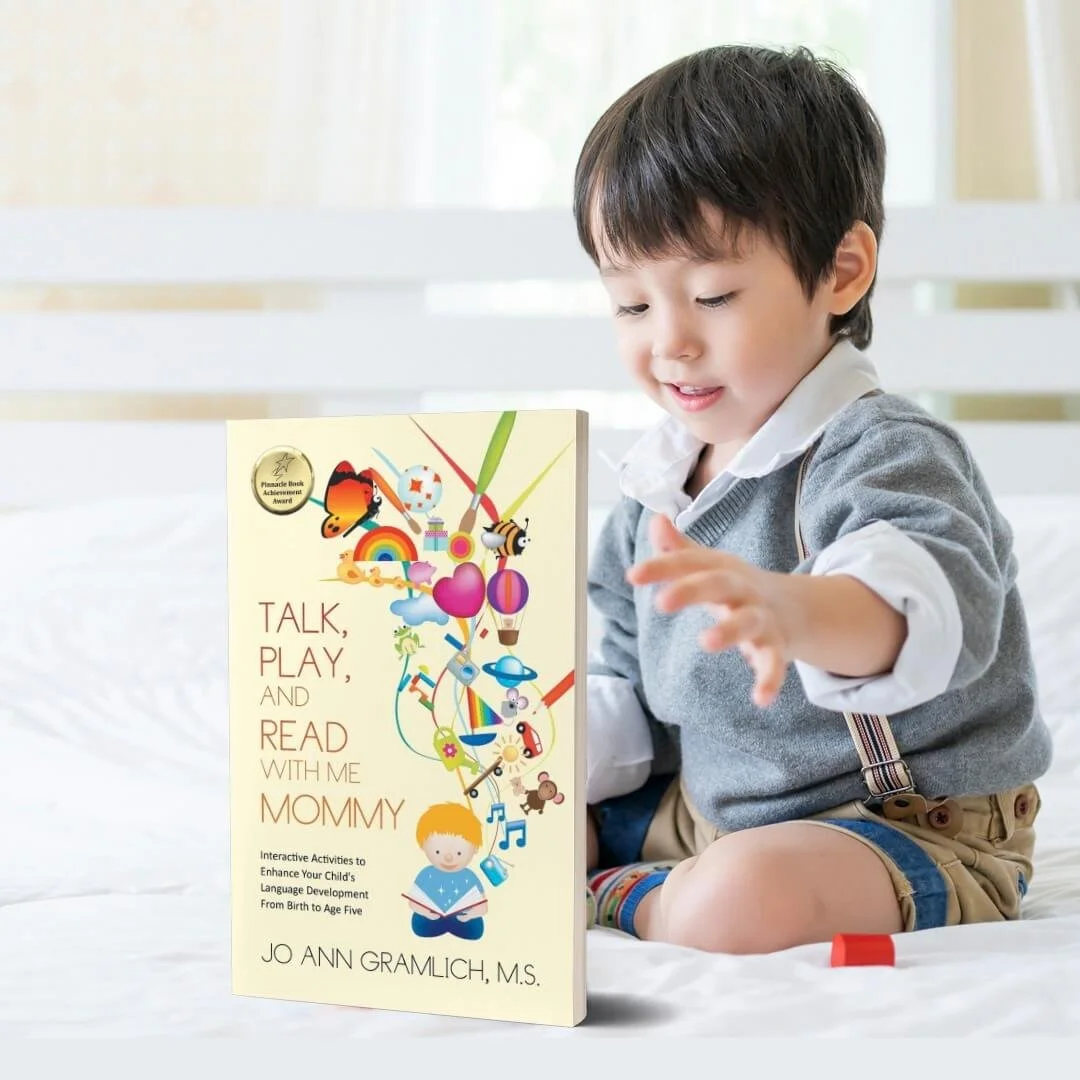 A young boy sitting on a white bed, reaching towards a book titled 'Talk, Play, and Read with Me Mommy.' The book cover features colorful illustrations of animals, rainbows, and playful objects.