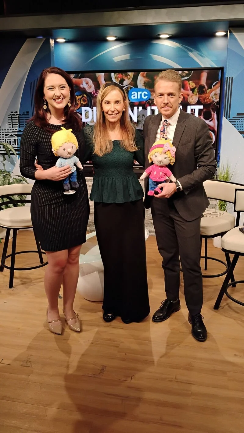 Three news anchors standing in a studio, each holding a plush doll, with a television screen behind them.